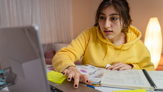 A student with a yellow hoodie sitting at their desk and working on their computer with a notebook and handout papers.