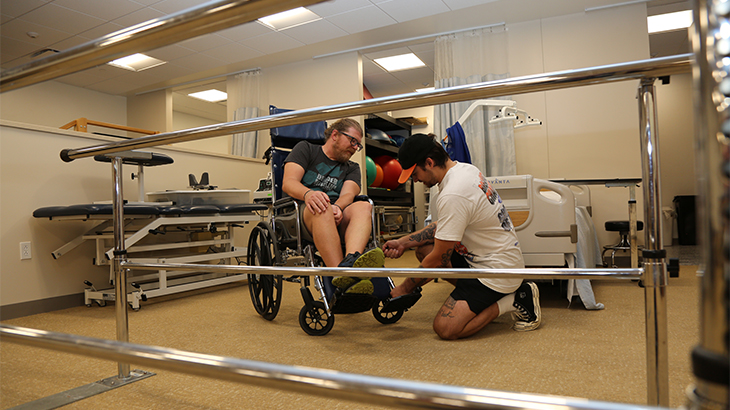 Image: photo of a PTA student sitting in a wheelchair while another student shows him how to use the wheelchair for patient injuries