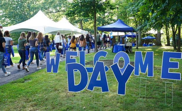 Welcome Day sign with students in the background walking on the paths outside