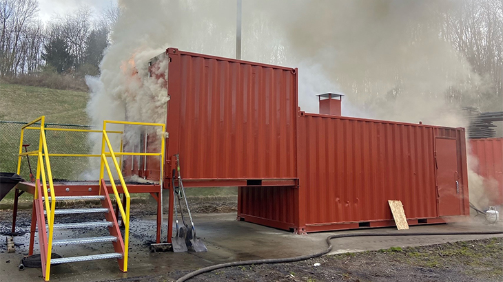 Image: photo of a simulated fire in a red shipping container with grey smoke billowing out of the top